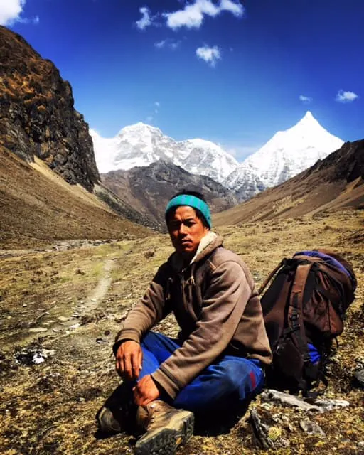 A man sitting on the ground with his backpack in front of mountains.