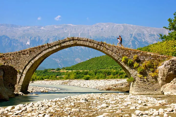 An old stone bridge over a river with mountains in the background.