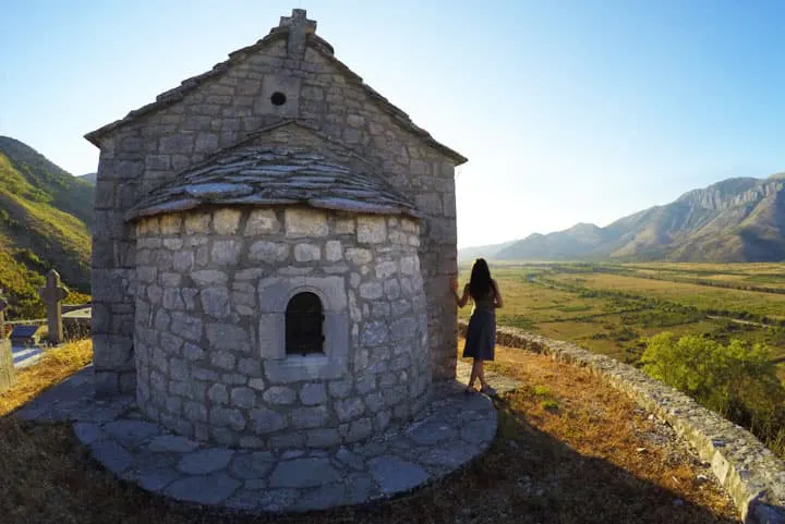 A woman is standing in front of a stone church in the mountains.