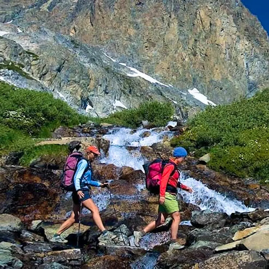 Two hikers walking along a stream with mountains in the background.
