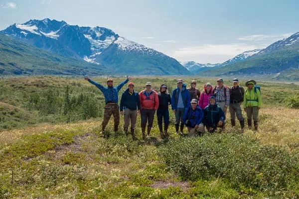 A group of people standing in a grassy area with mountains in the background.