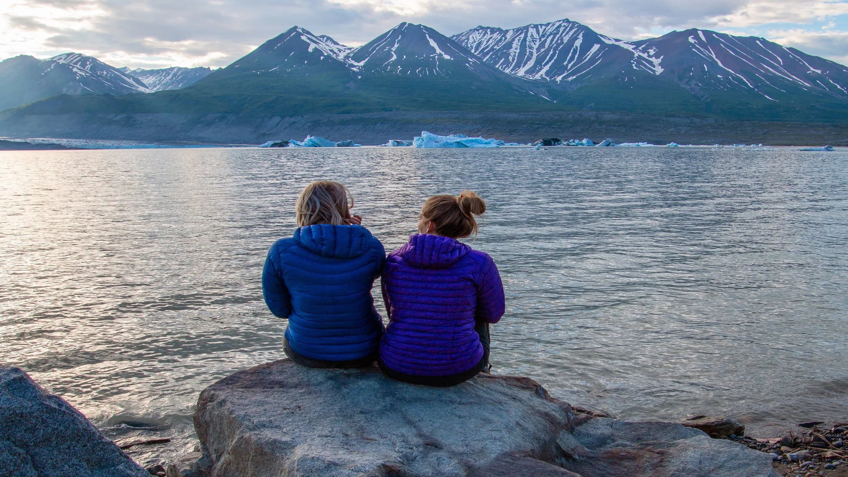 Two women sitting on a rock in front of a body of water.