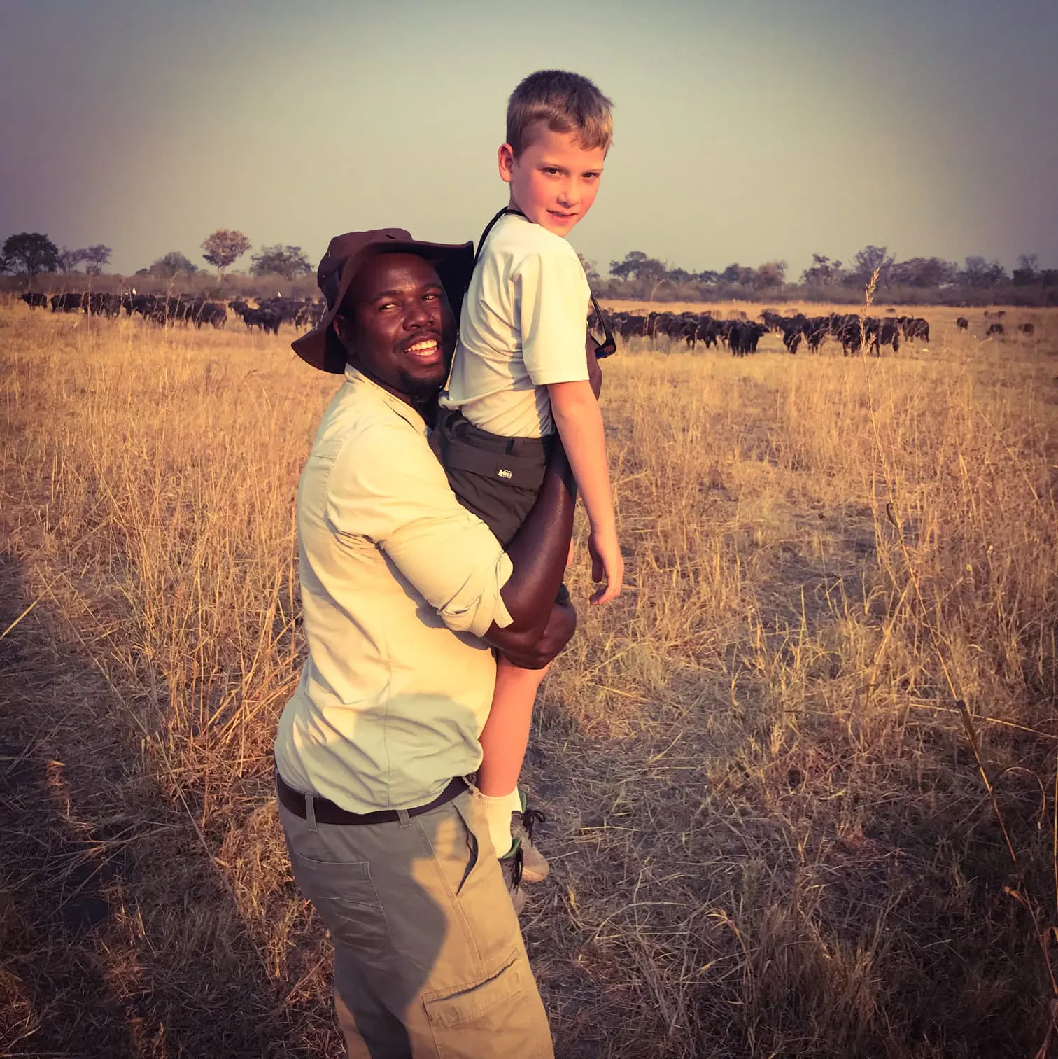 A man holding a young boy in front of a herd of wildebeest.