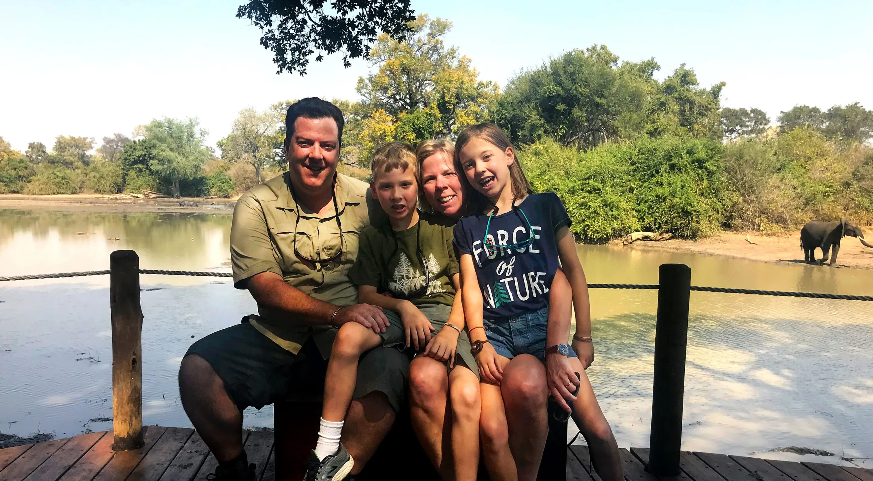 A family is posing for a photo on a wooden deck with an elephant in the background.