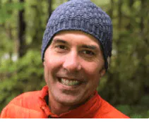 A man in an orange jacket smiling in the woods.