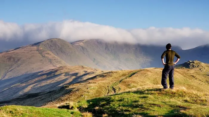 A man standing on a grassy hill overlooking mountains.