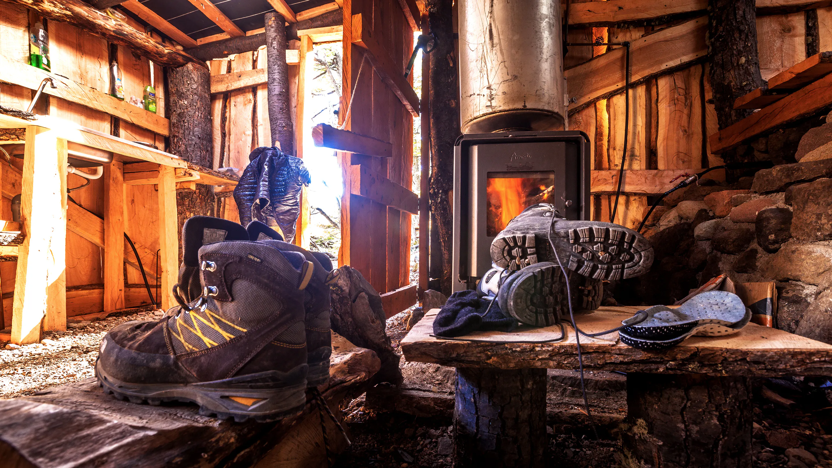 A pair of boots sitting on a wooden bench next to a wood burning stove.
