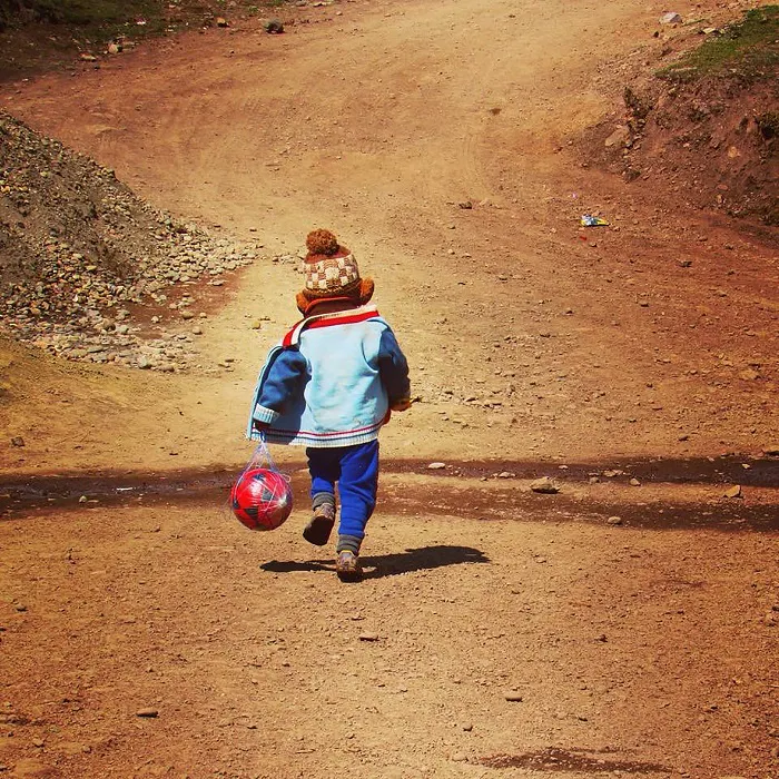 A child is running down a dirt road with a ball.
