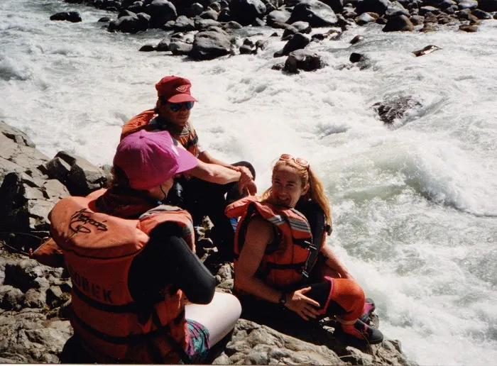 Three people in life jackets sitting on a rock next to a river.