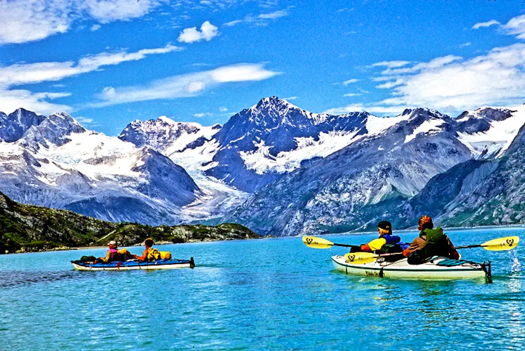 A group of people are kayaking in the water with mountains in the background.