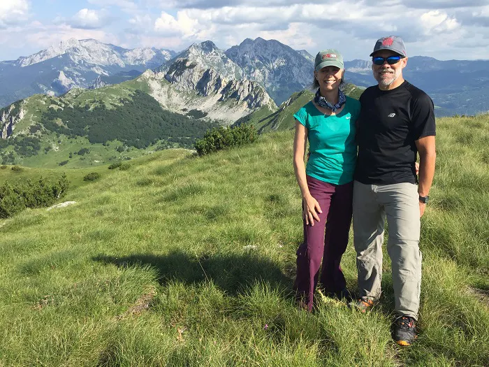 A man and woman standing on a grassy hill with mountains in the background.