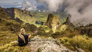 A woman sits on top of a mountain overlooking the clouds.