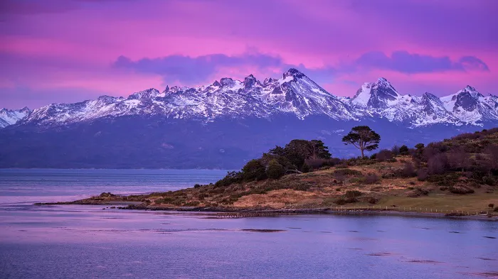 A purple sky over a lake with snow capped mountains in the background.