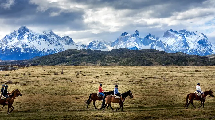 A group of people riding horses in a field with mountains in the background.