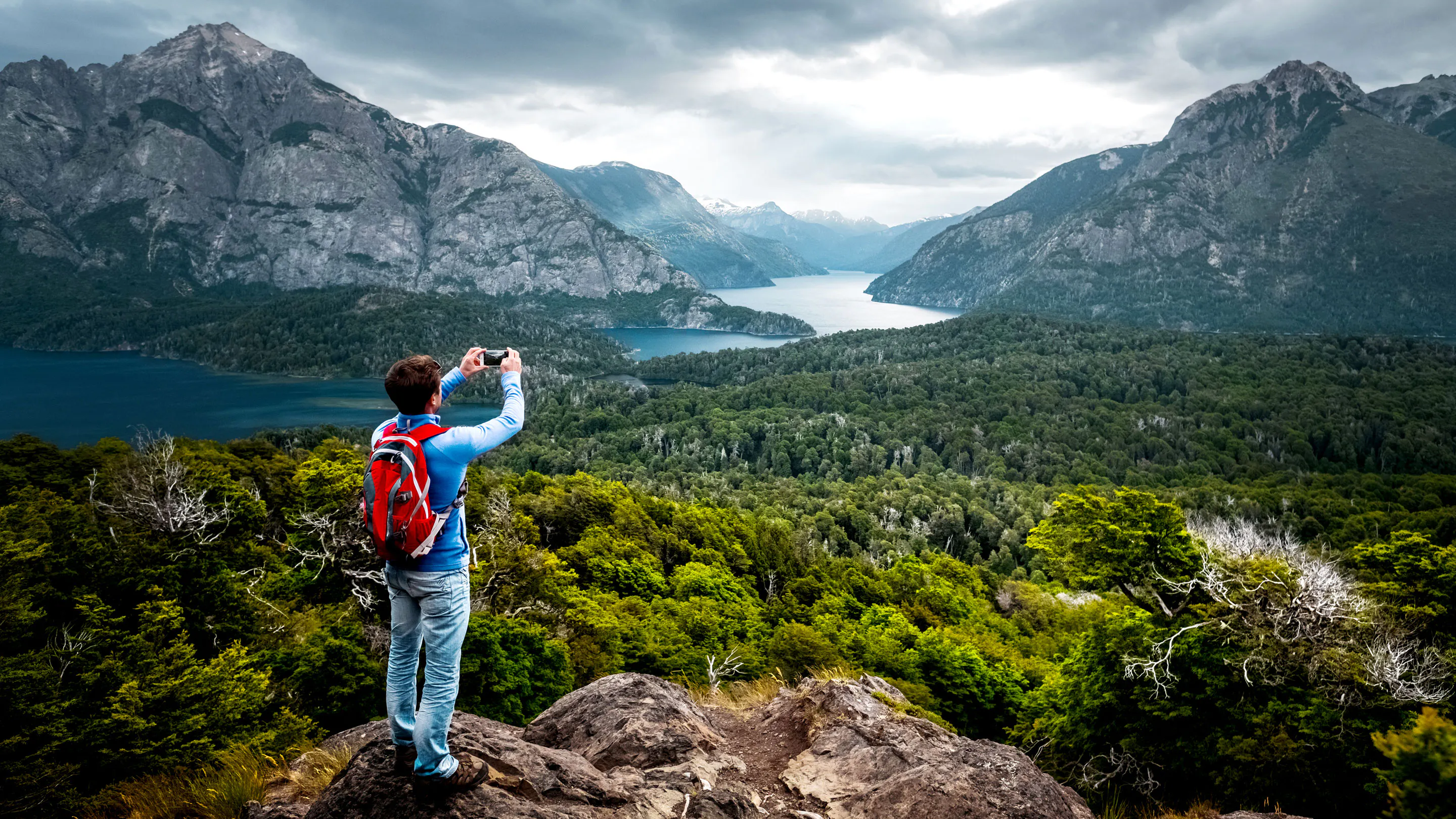 A man is taking a picture of a lake and mountains.
