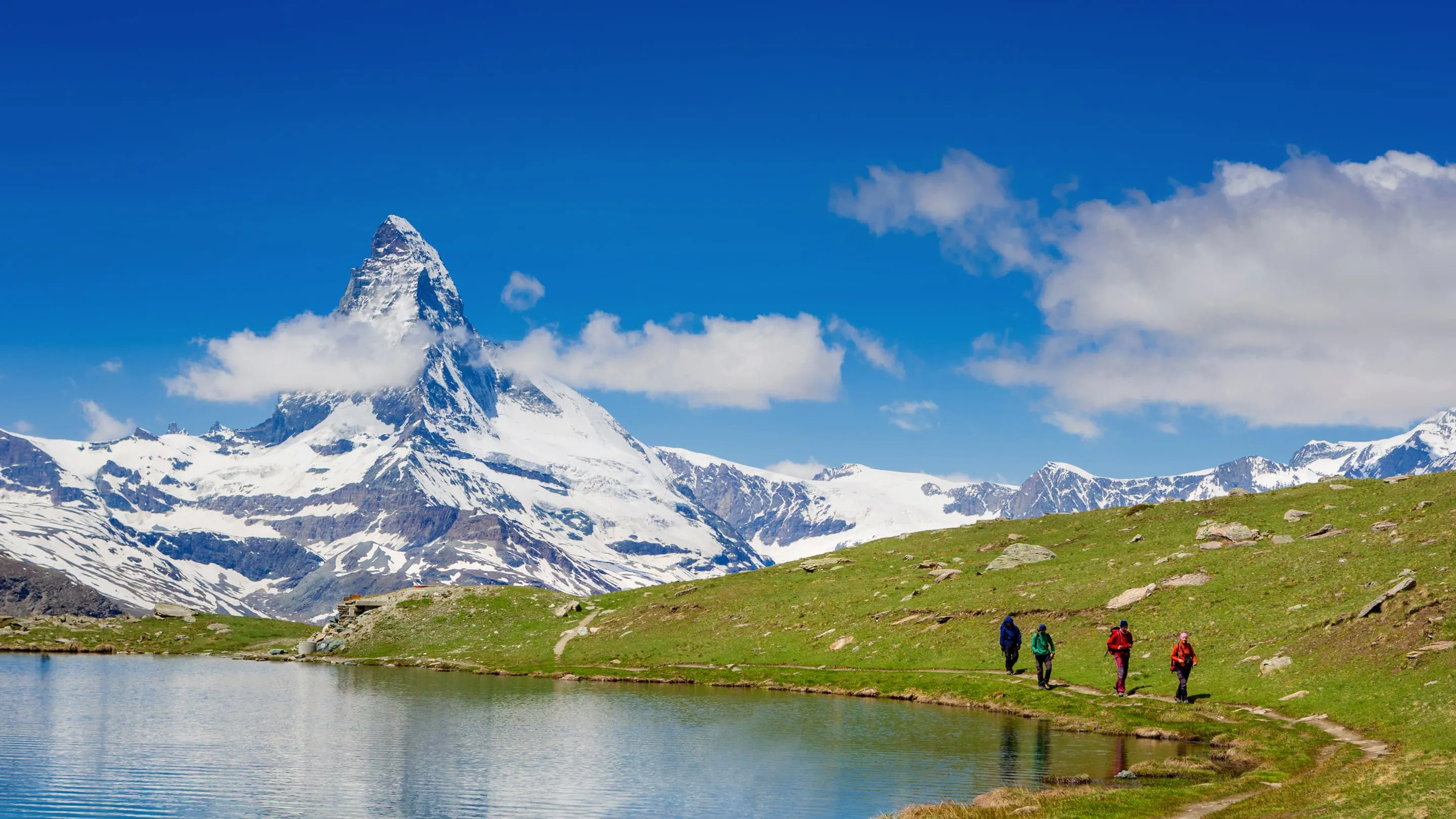 A group of people walking near a lake with a mountain in the background.