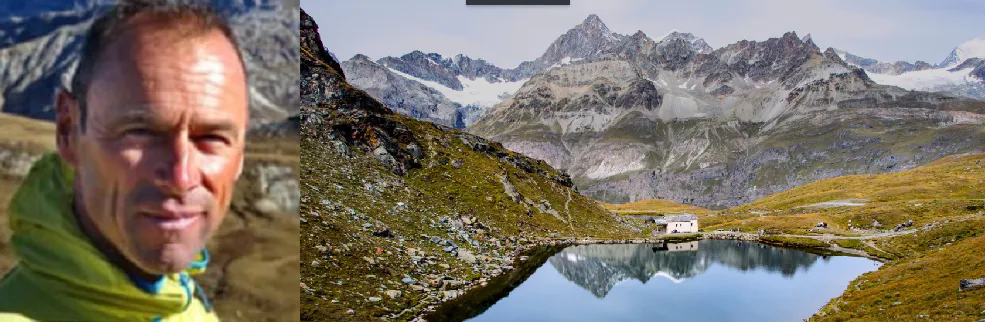 A man is standing in front of a mountain with a lake in the background.