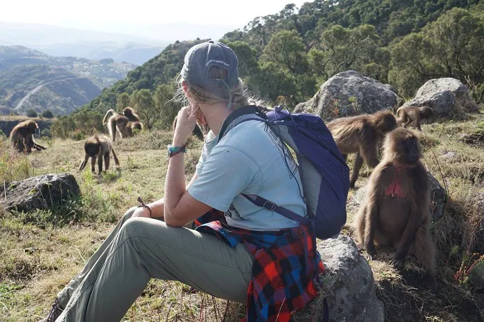 A woman sits on a rock with baboons in the background.