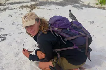 A woman kneeling on the sand with a backpack and a bird on her shoulder.
