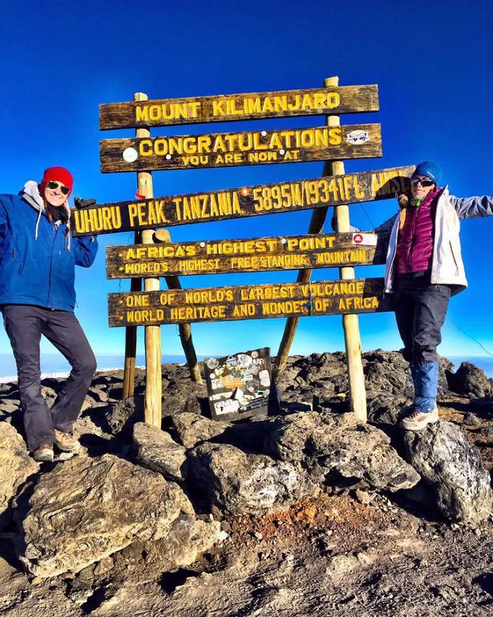 Two people standing in front of a wooden sign on top of a mountain.