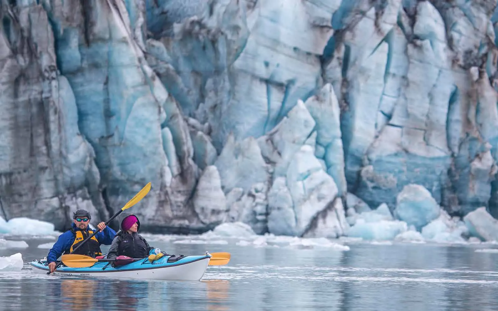 Two people paddling in a kayak in front of an iceberg.