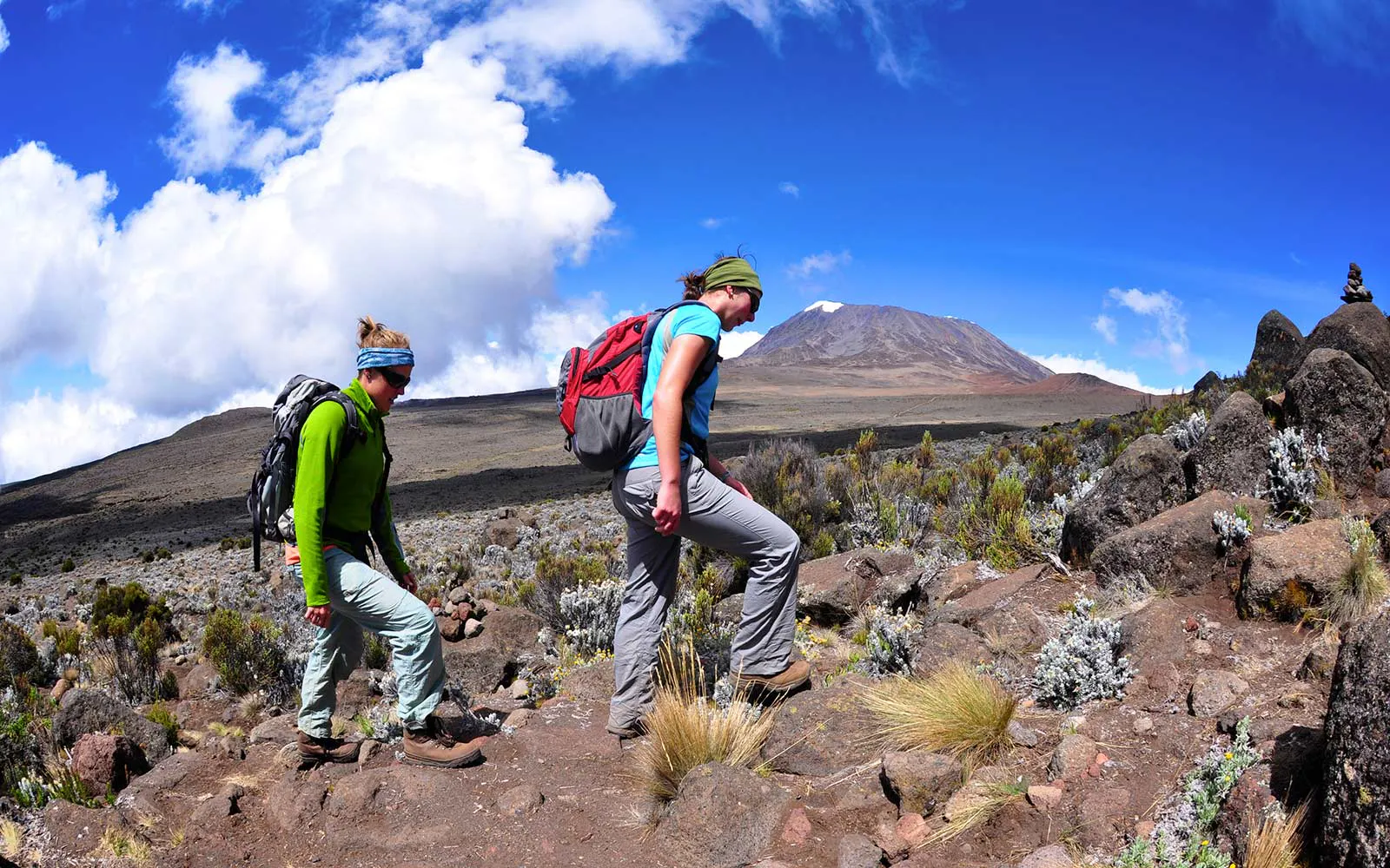 Two people embarking on a challenging hike along a rugged trail with a majestic mountain towering in the background.