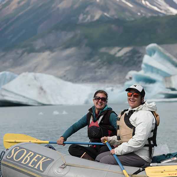Two people in a raft with icebergs in the background.