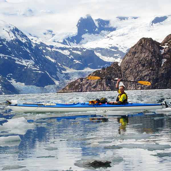 A man paddles a kayak in a body of water with icebergs in the background.