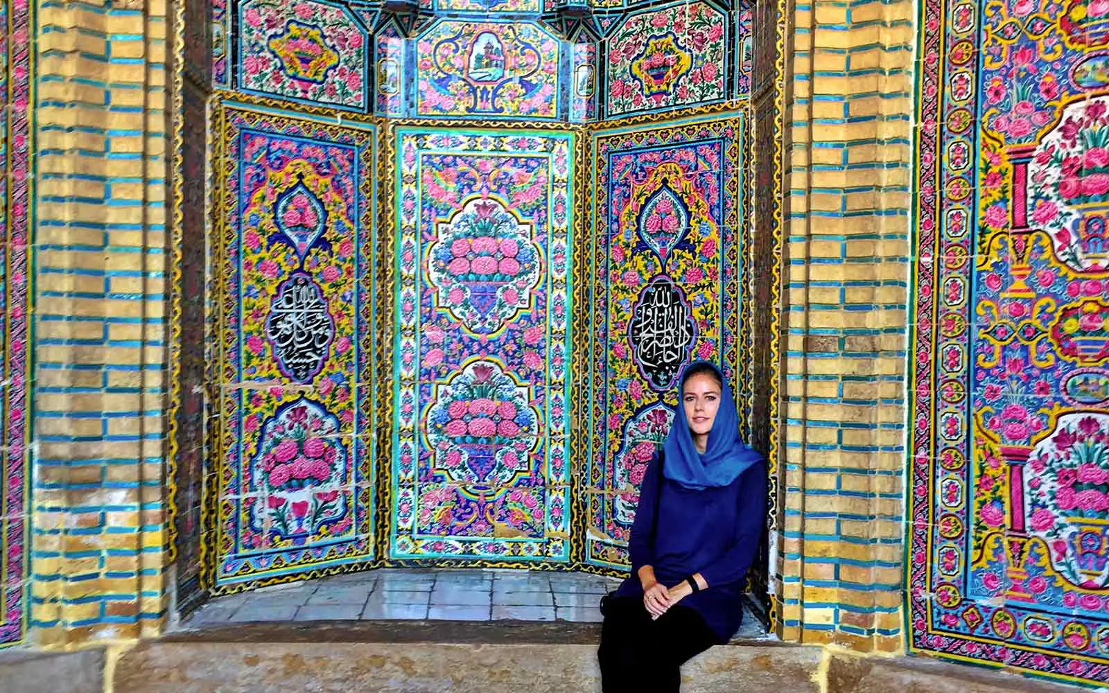 A woman in a hijab sitting in front of colorful tiled walls.