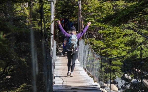 A woman is crossing a suspension bridge with her arms up, experiencing one of the best adventures in Patagonia.