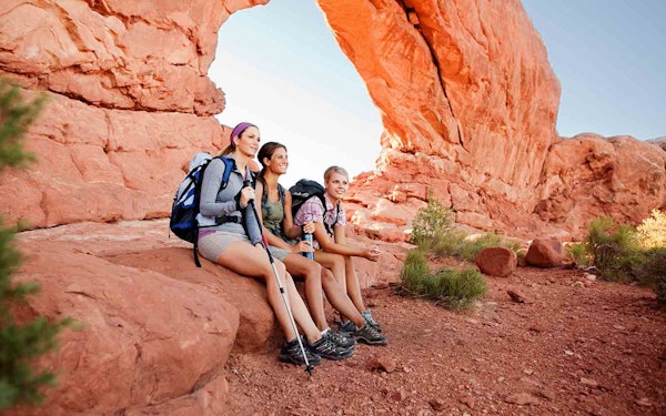 Three hikers enjoying national park adventures while sitting in front of a breathtaking red rock arch.