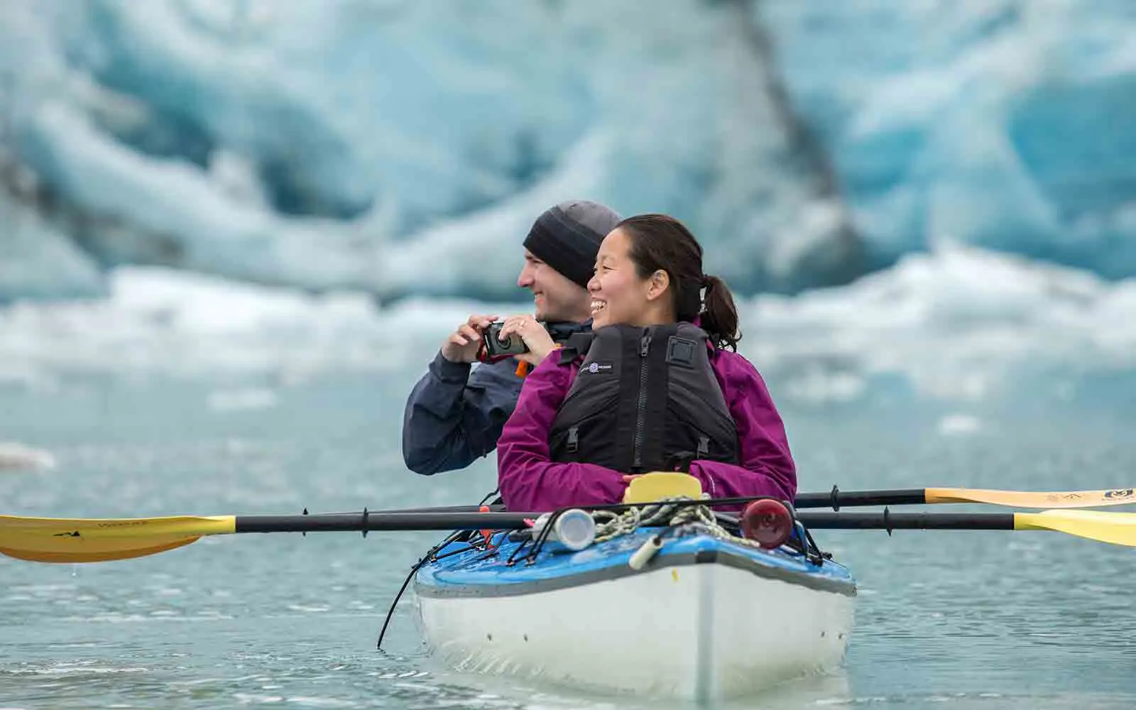 Two people enjoying national park adventures while paddling a kayak in front of an iceberg.