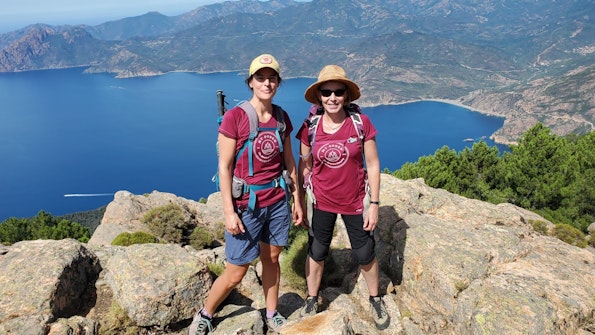 Two women on a thrilling adventure travel, standing on top of a mountain with a breathtaking view of the sea.