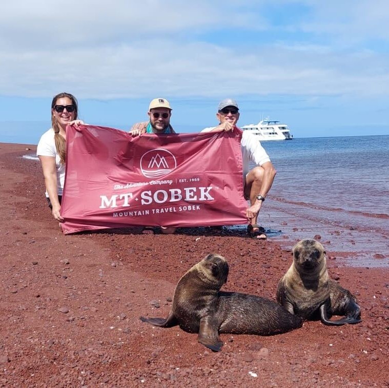 Three people standing on a beach with seals and a sign that says Mr. Sobek, engaging in adventure travel.