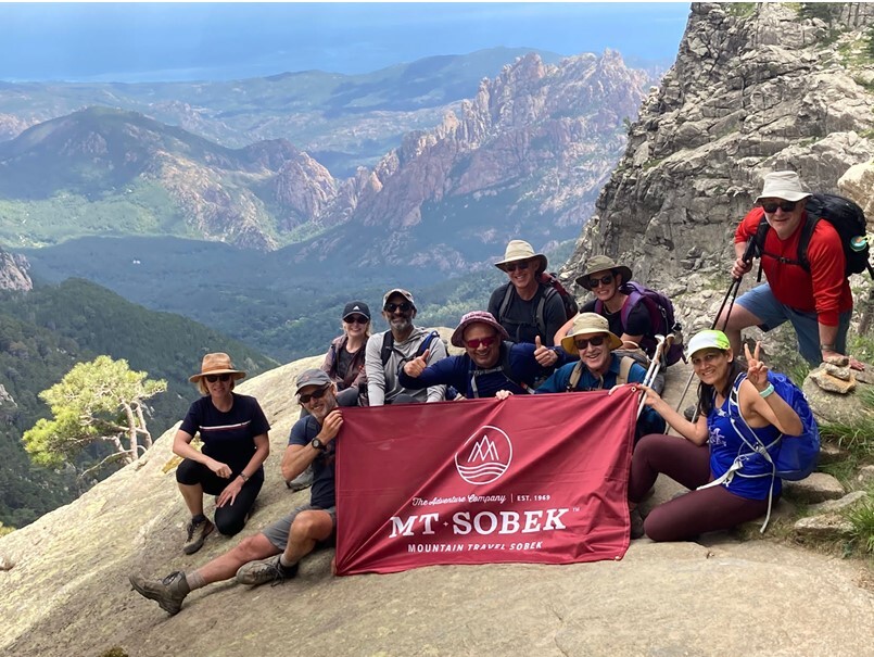 A group of people on an adventure travel posing on a mountain with a banner.