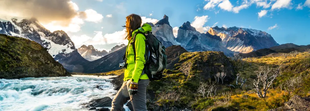 A hiker standing in front of a river with mountains in the background.