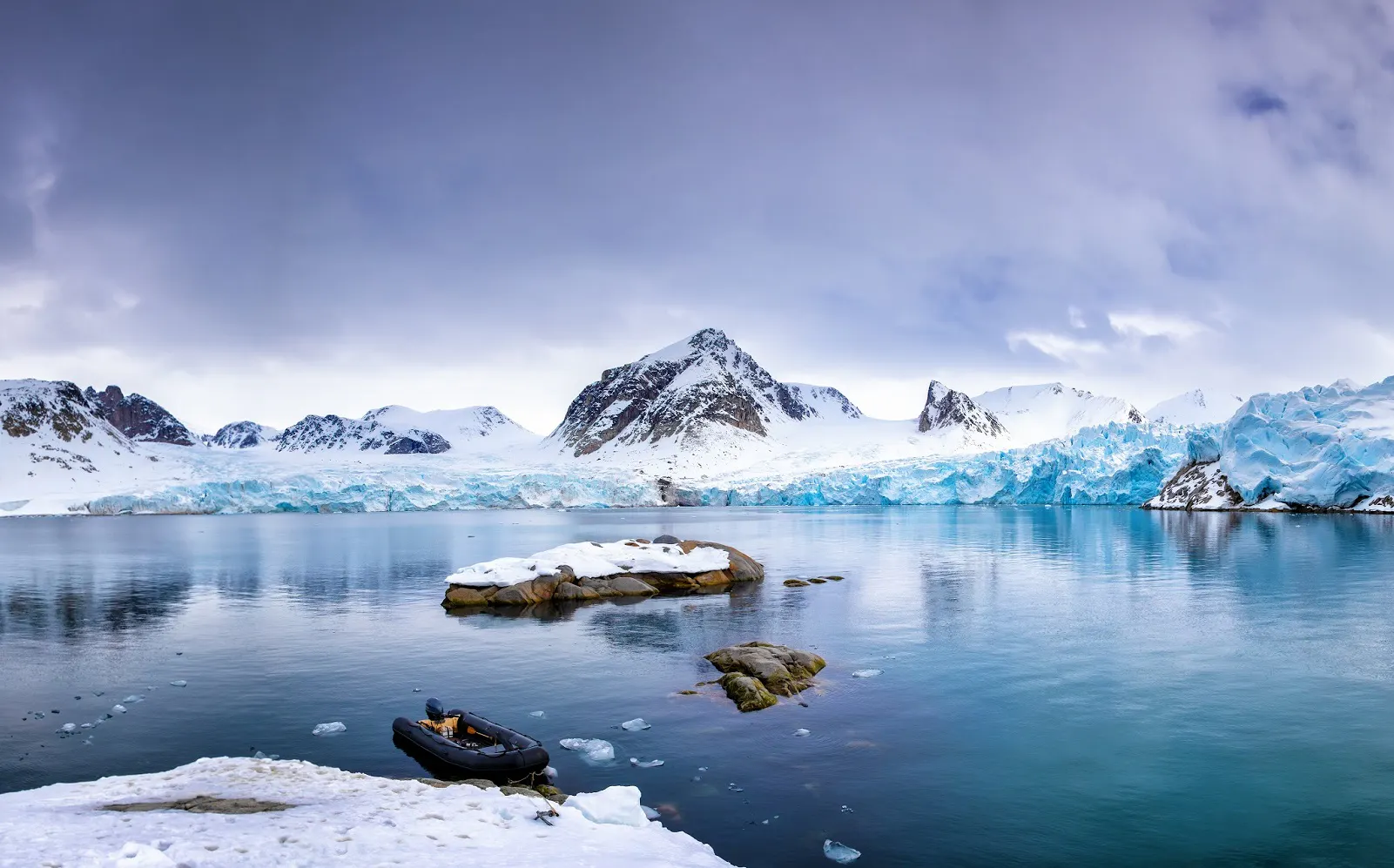 Snow-covered mountains and glaciers border a calm, icy body of water, with a small inflatable boat moored near rocks in the foreground under a cloudy sky.