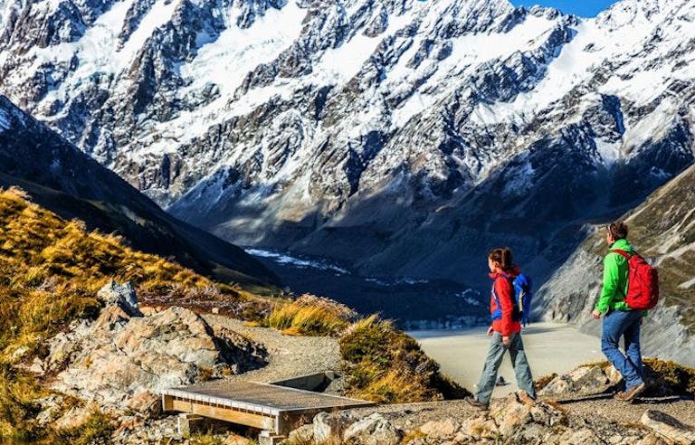 Two hikers on a trail with rugged, snowy mountains in the background in New Zealand.