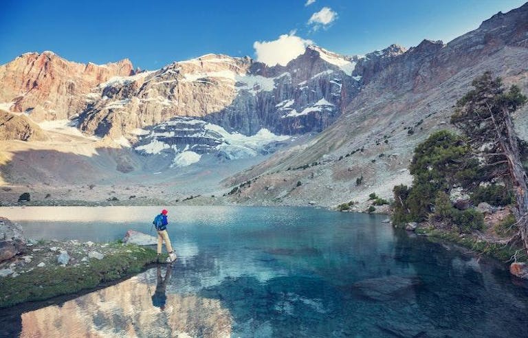 Man hiking in the Tajikistan Fann Mountain range with a crystal clear river and snowy mountains in the background