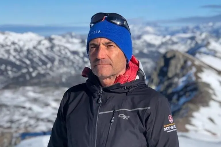 A man wearing a blue beanie, sunglasses, and a black jacket stands outdoors with snow-covered mountains in the background.