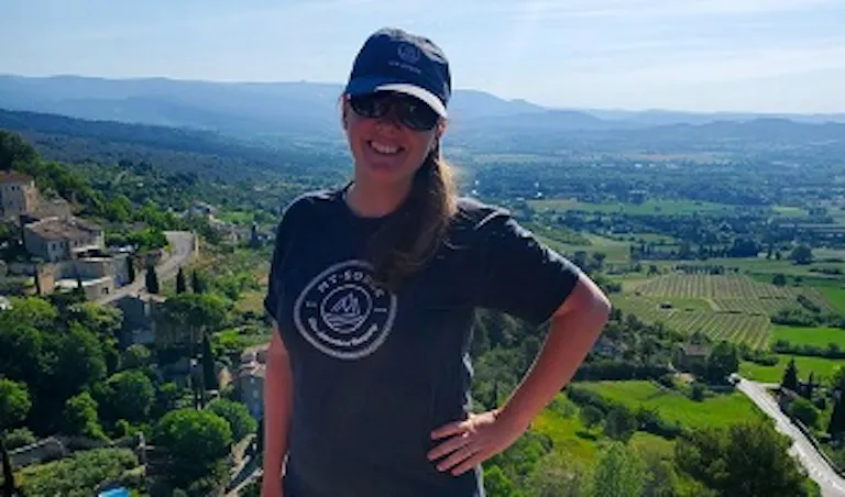 Woman wearing sunglasses, a hat, and a dark t-shirt stands outdoors with a scenic view of hills, trees, and fields in the background under a clear sky.