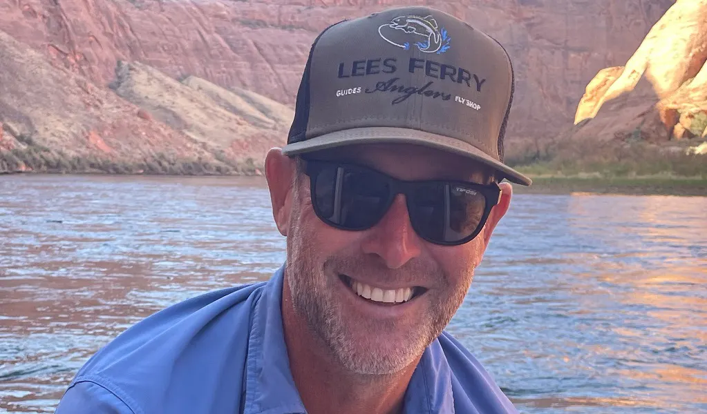 Man wearing sunglasses and a “Lees Ferry Anglers” hat smiles by a river with rocky cliffs in the background.