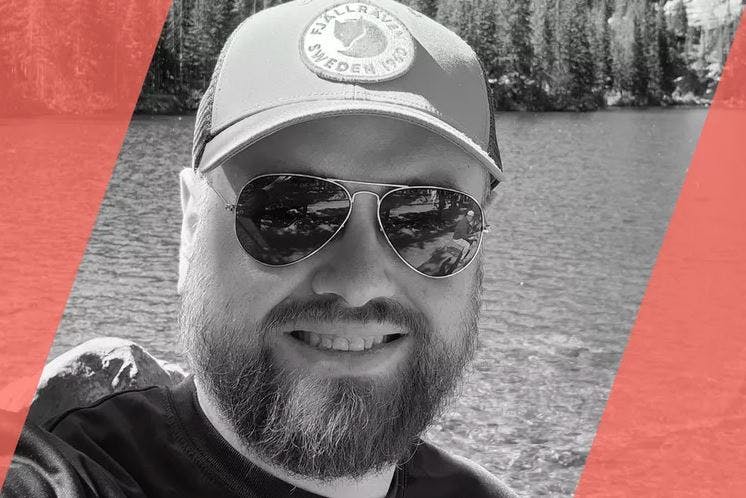 A travel advisor with a beard and sunglasses is taking a selfie in front of a lake.