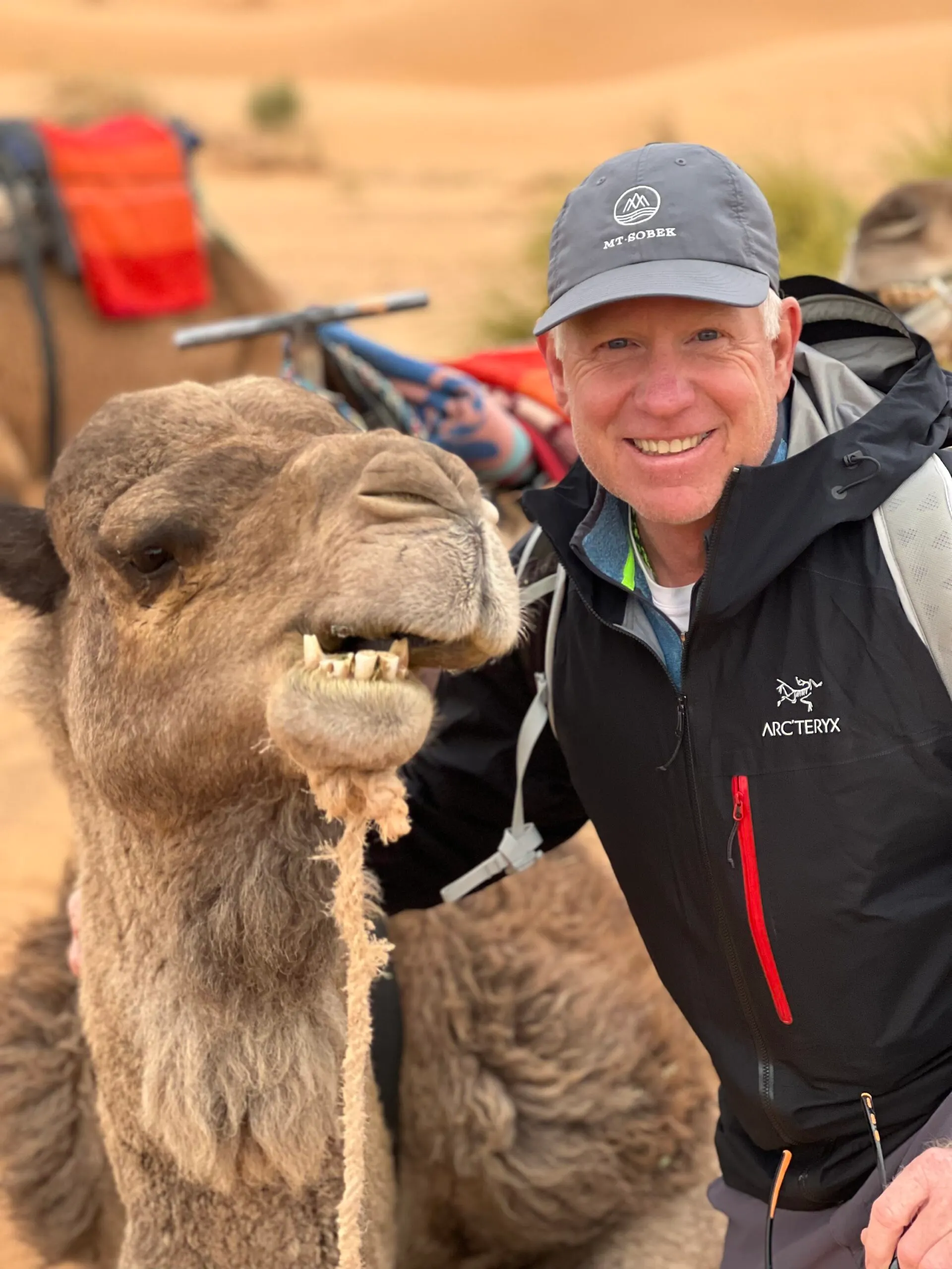 A man smiling next to a camel in the desert, entered in a photo contest.