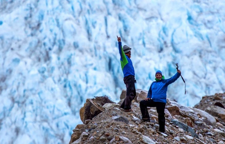 two males in warm winter gear and trekking poles on top of rock overlooking large Patagonia glacier