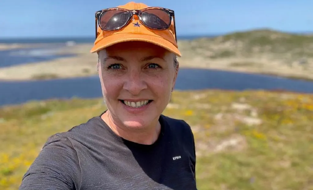 Woman wearing an orange cap and sunglasses takes a selfie outdoors with a grassy landscape, water, and blue sky in the background.