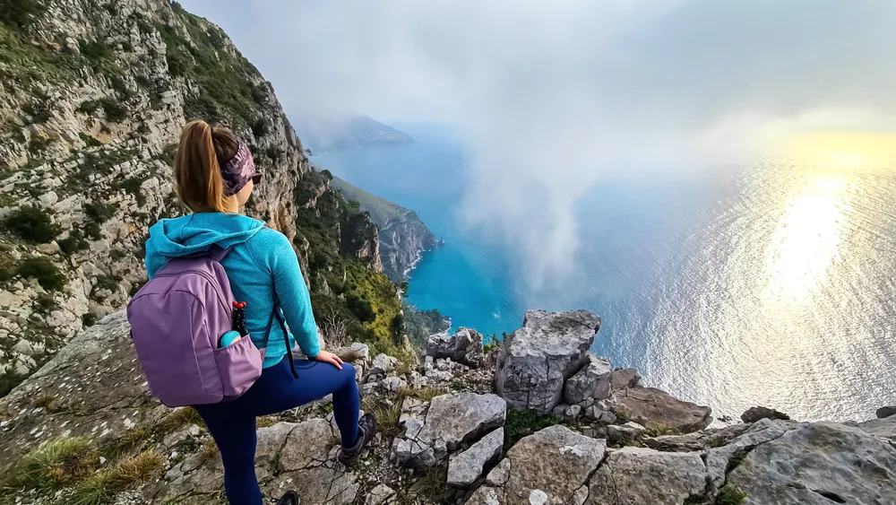 Woman with scenic view from a hiking trail on the coastal driving road of beautiful Amalfi Coast, Campania, Italy, Europe. Riviera coastline at Mediterranean sea. Panoramic curvy road near Positano
