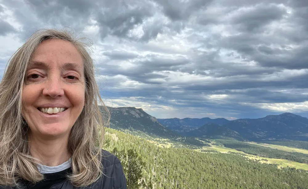 A woman with gray hair smiles at the camera in front of a scenic mountain landscape with cloudy skies.