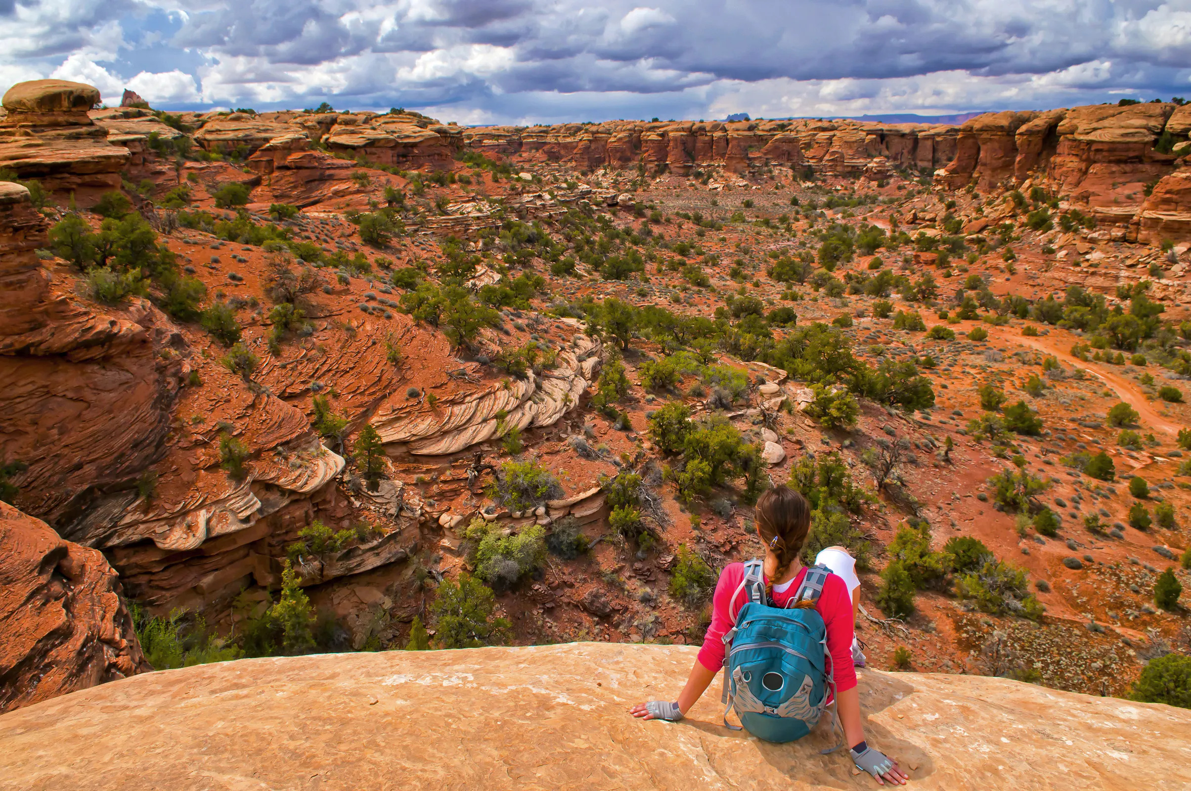 A person with a teal backpack sits on a rocky ledge overlooking a wide canyon landscape with layered red rock formations and scattered shrubs under a cloudy sky.