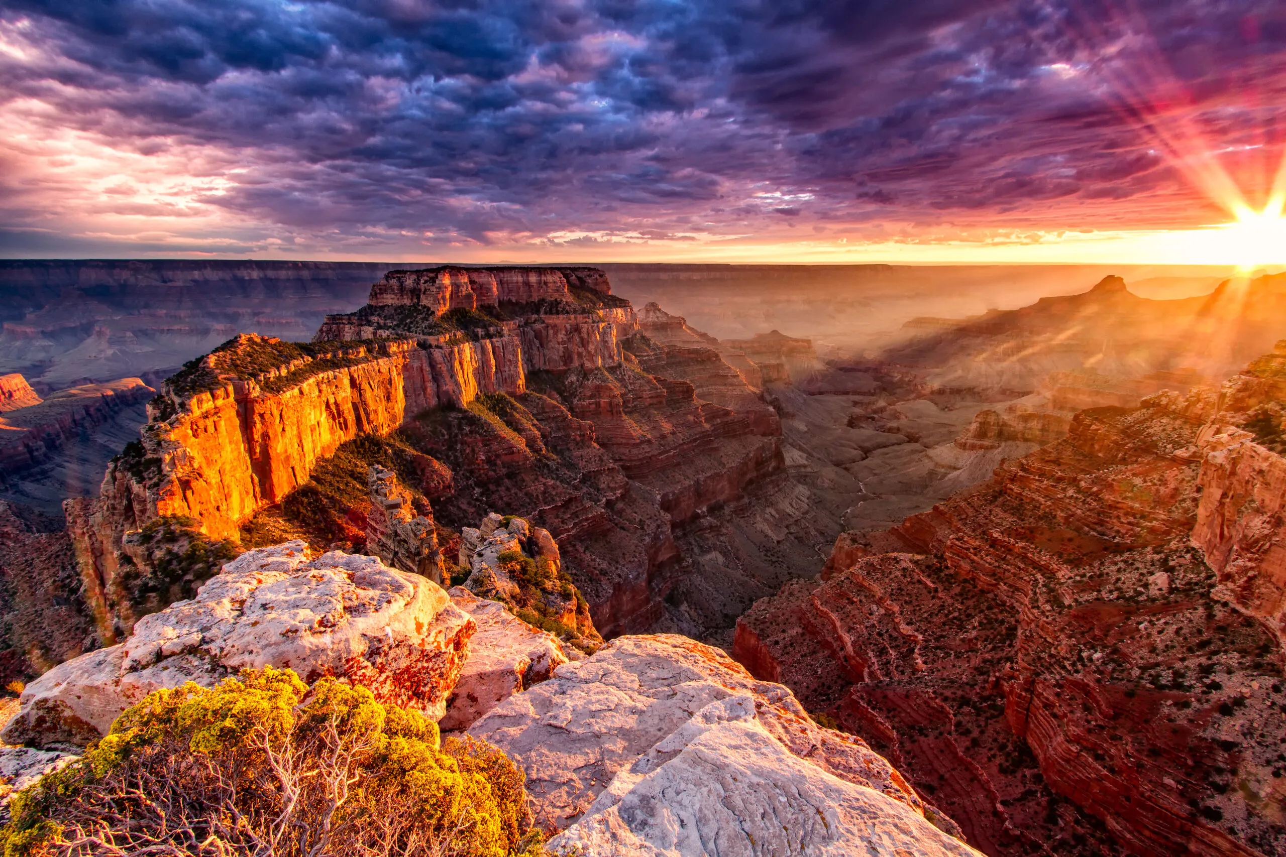 Sunrise over the Grand Canyon, with sunlight illuminating layered rock formations and dramatic clouds in the sky.